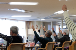 介護施設での入居者によるレクリエーション活動。複数の高齢者が腕を上げて体操や運動に参加している。